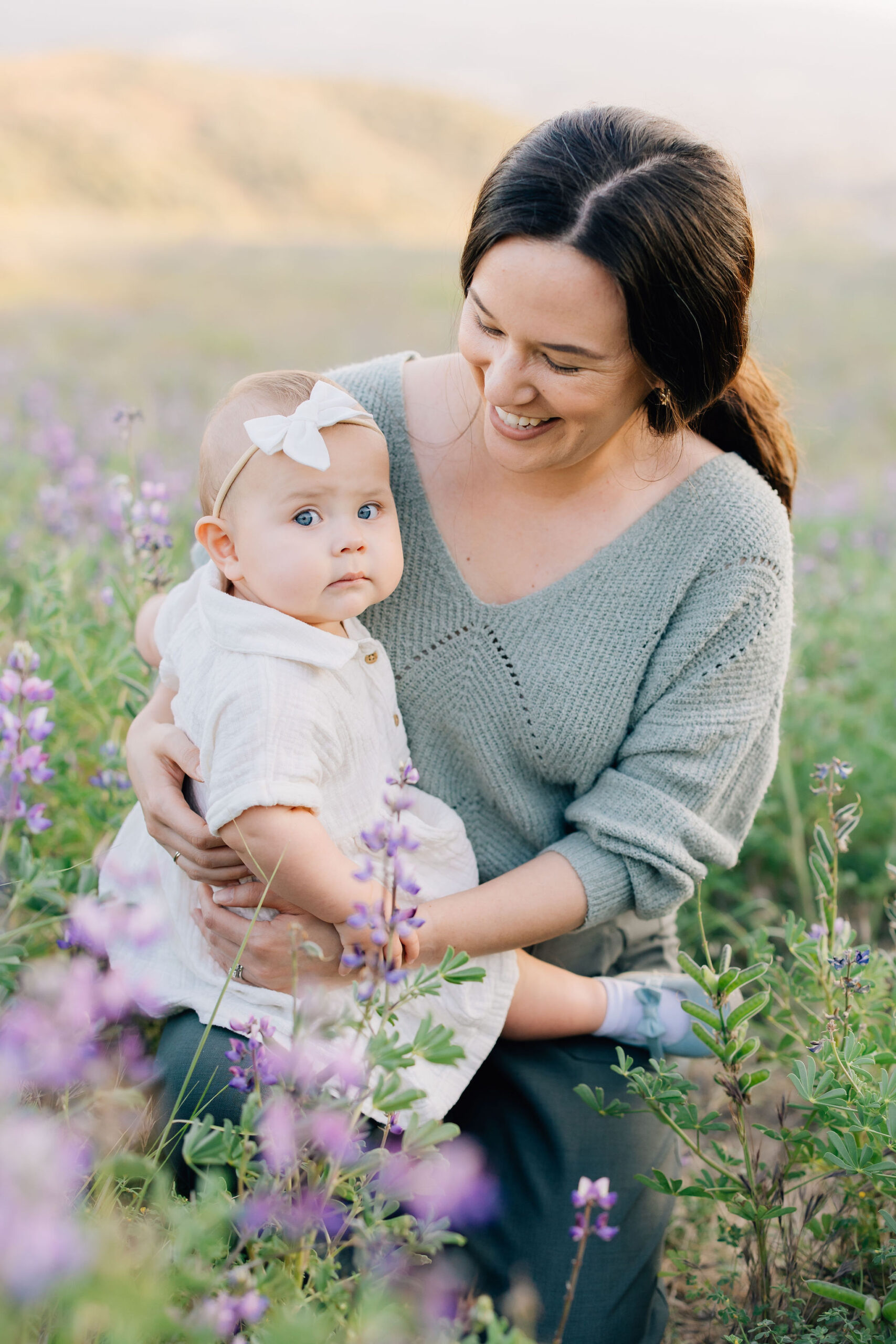 A happy mom sits in a valley of purple flowers with her toddler daughter in a white dress in her lap