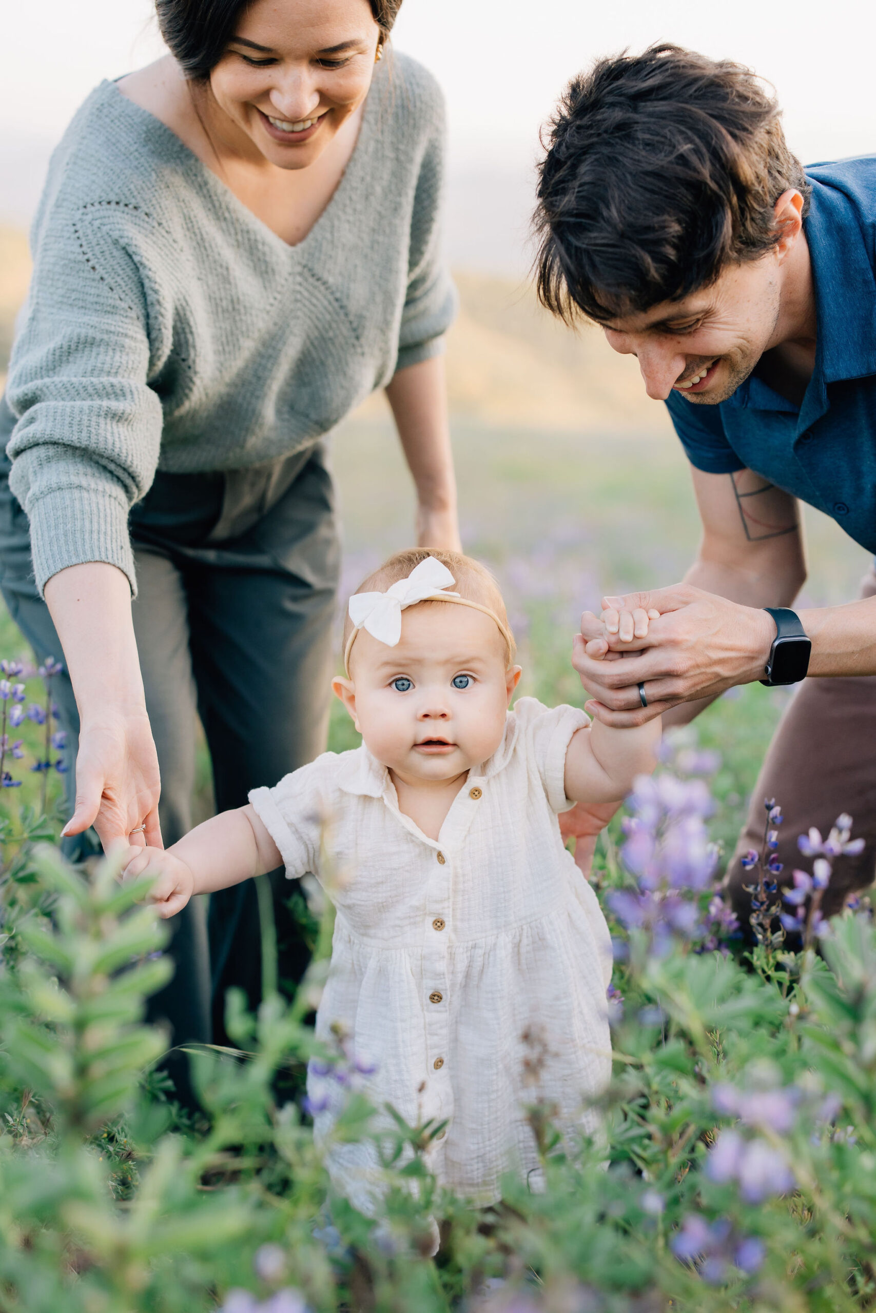 A toddler girl in a white dress and bow explores wildflowers with help from mom and dad before visiting baby stores in santa clarita ca