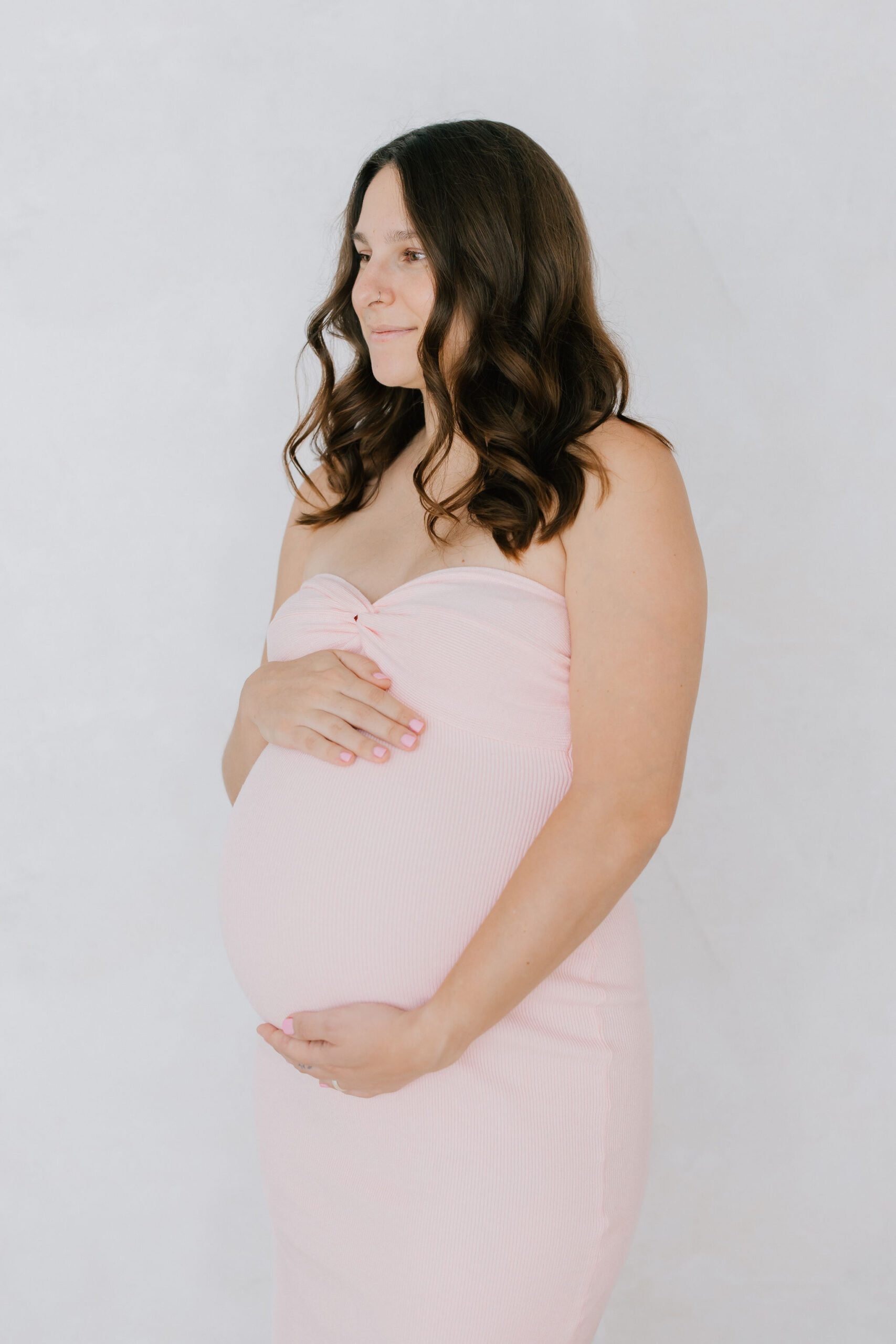 A smiling mom to be in a pink maternity gown stands in a studio holding her bump