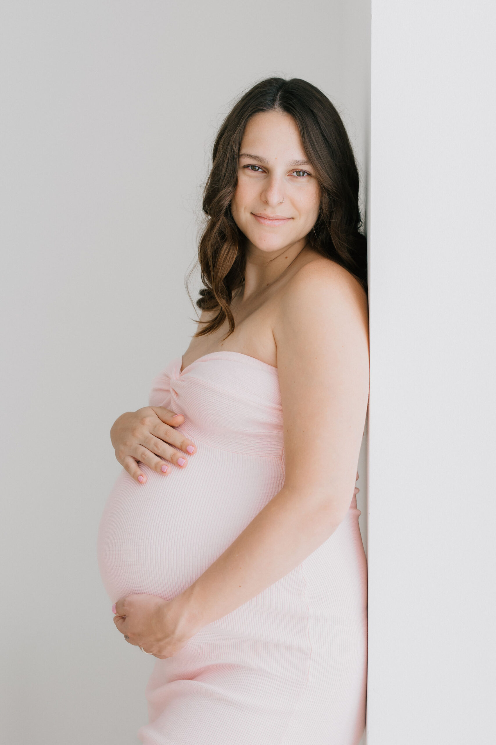 A pregnant woman leans on a wall in a pink maternity gown while smiling down her shoulder after visiting a birthing center santa clarita