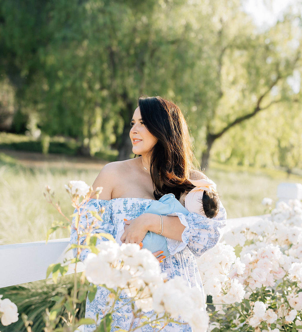 A mom explores a garden in a blue dress while carrying her sleeping newborn baby