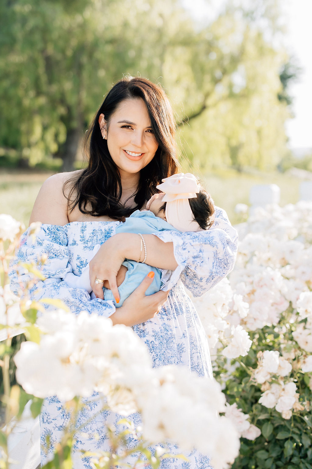 A smiling mom stands in a garden holding her sleeping newborn baby among white flowers after meeting a doula santa clarita