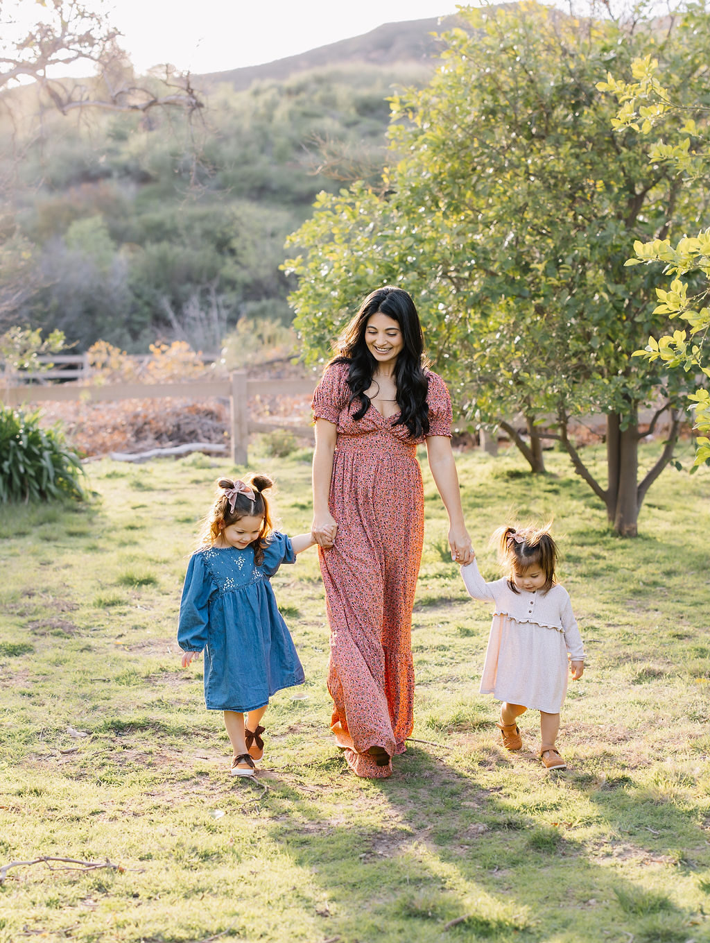 toddler sisters hold hands with mom while exploring a park at sunset