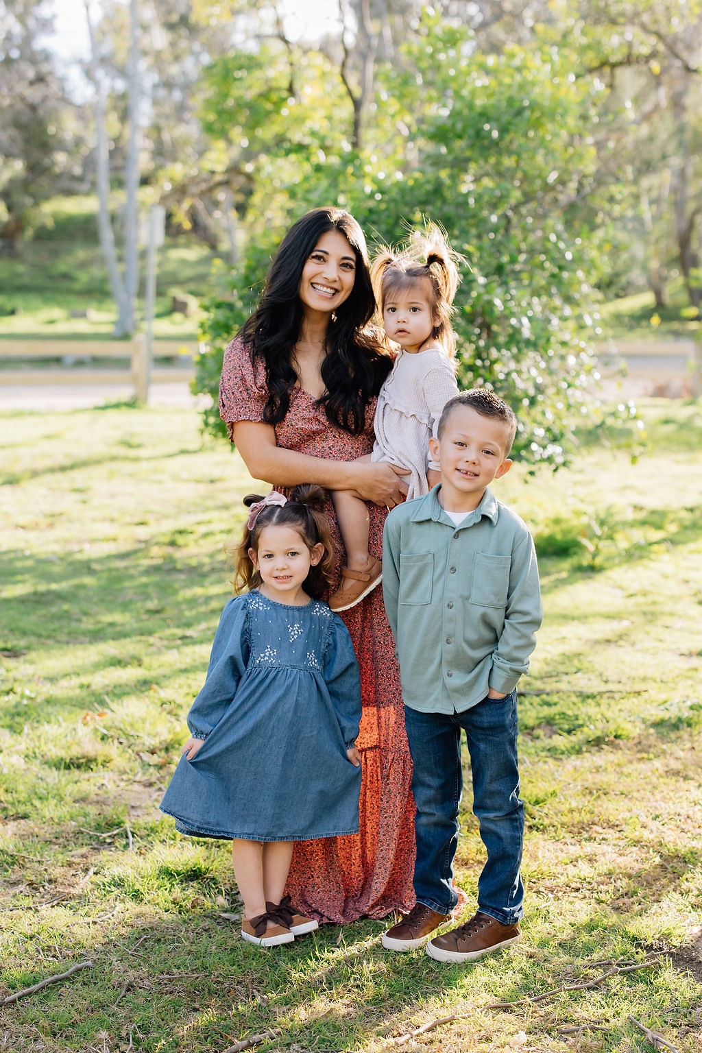 A smiling mother stands in a park with her 2 toddler daughters and toddler son before visiting pumpkin patches santa clarita