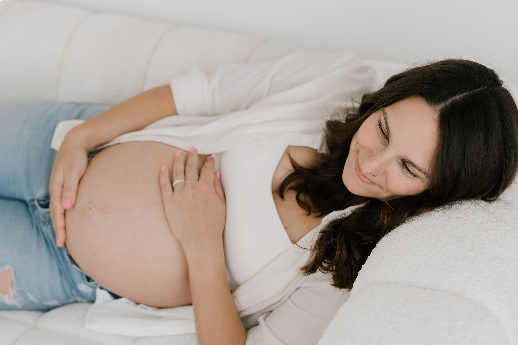 A smiling pregnant woman in an open white shit and jeans lays across a couch holding her bump