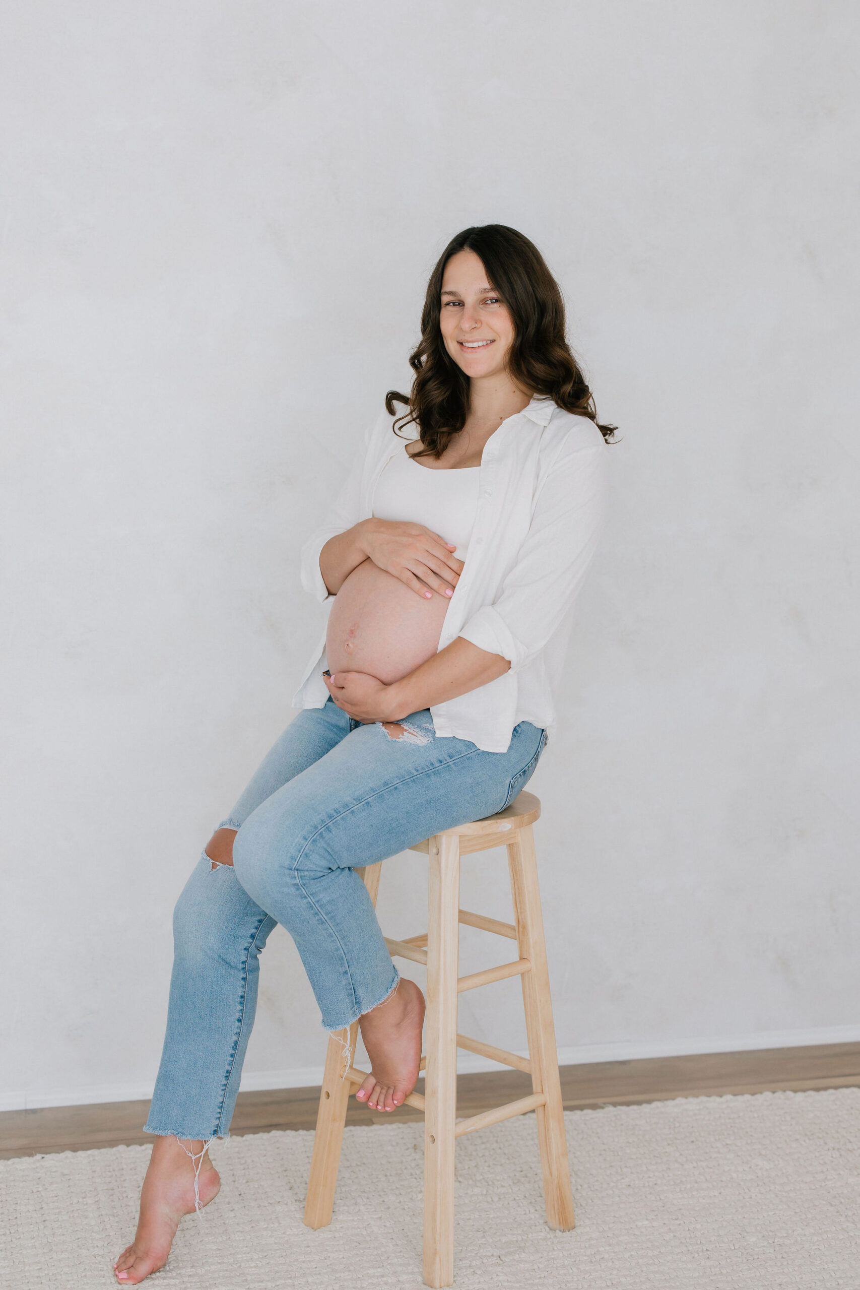 A happy mom to be sits on a wooden stool holding her exposed bump after a 3d ultrasound santa clarita