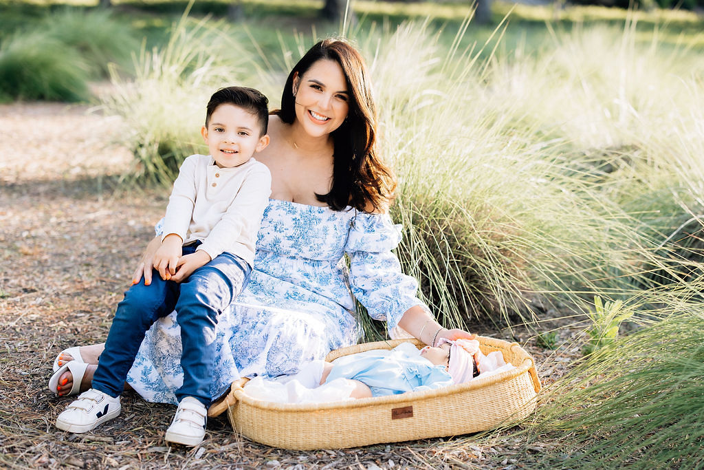 A happy mother sits in a park with her toddler son in her lap and newborn daughter laying in a wicker basket