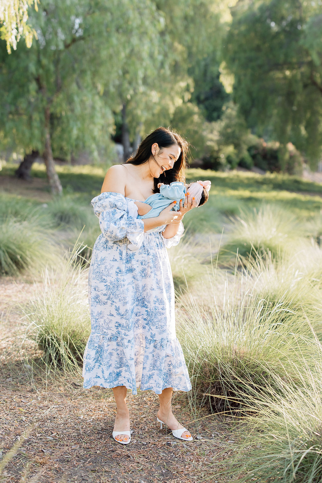 A happy mom in a blue floral print dress giggles with her newborn baby in a park of grasses at sunset before visiting daycares santa clarita