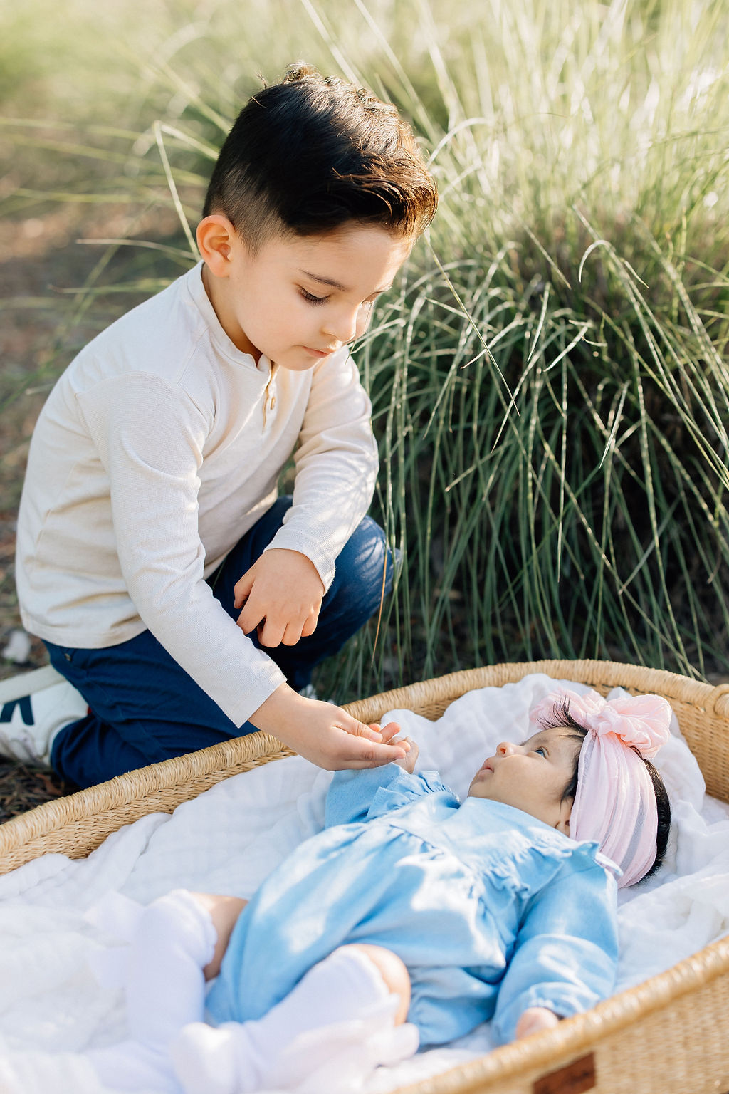 A toddler boy kneels over his newborn baby sister in a wicker basket while holding her hand before visiting daycares santa clarita