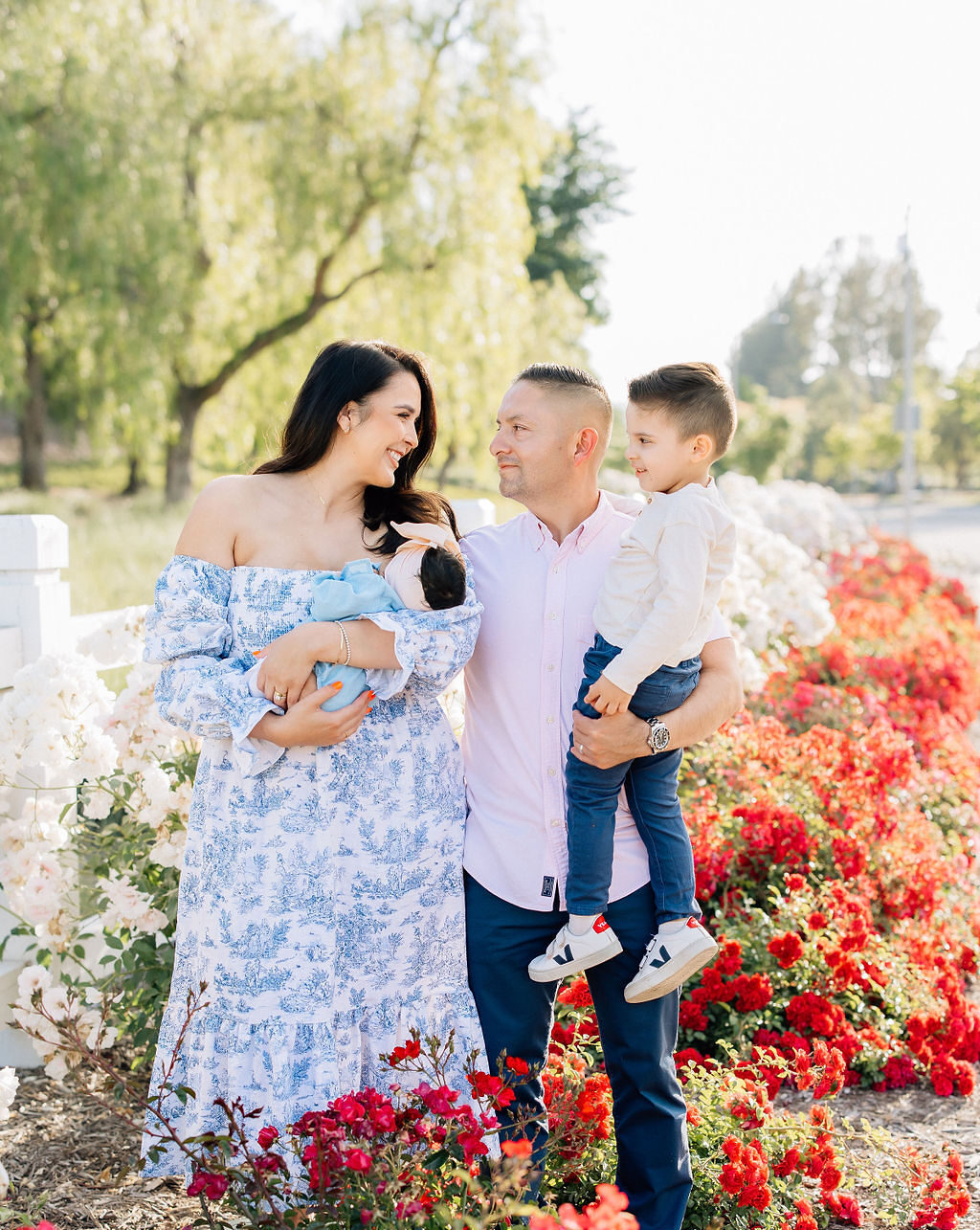 A happy mom holds her newborn baby in a blue dress while standing in a flower garden with dad holding their toddler son