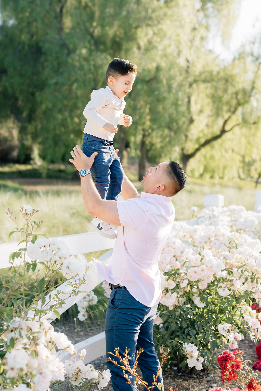 A dad tosses his giggling toddler son in matching shirt and pants while in a garden at sunset before visiting fall festival santa clarita