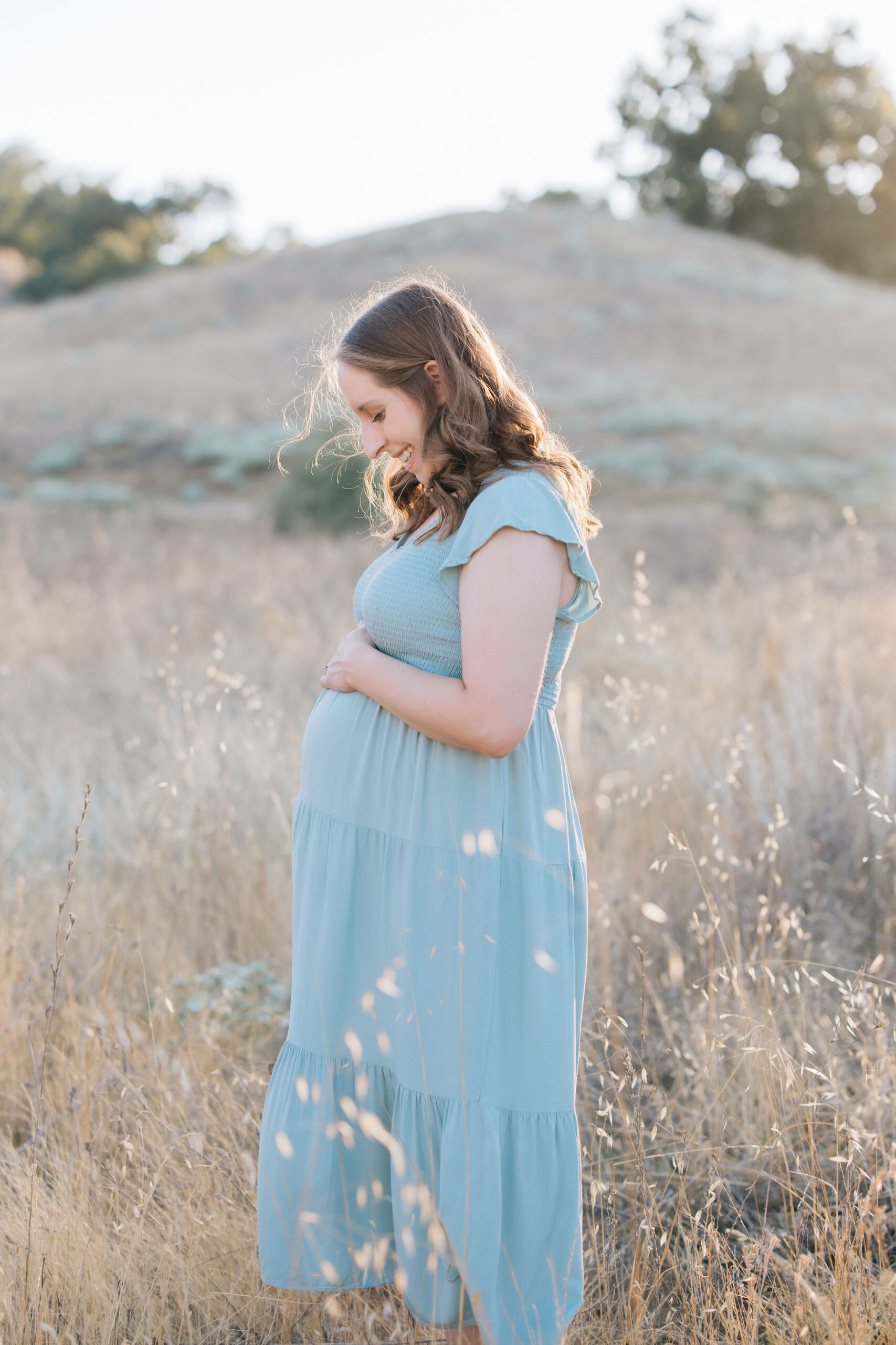 A happy mother to be smiles down to her bump in a blue maternity gown in a field of tall grasses at sunset