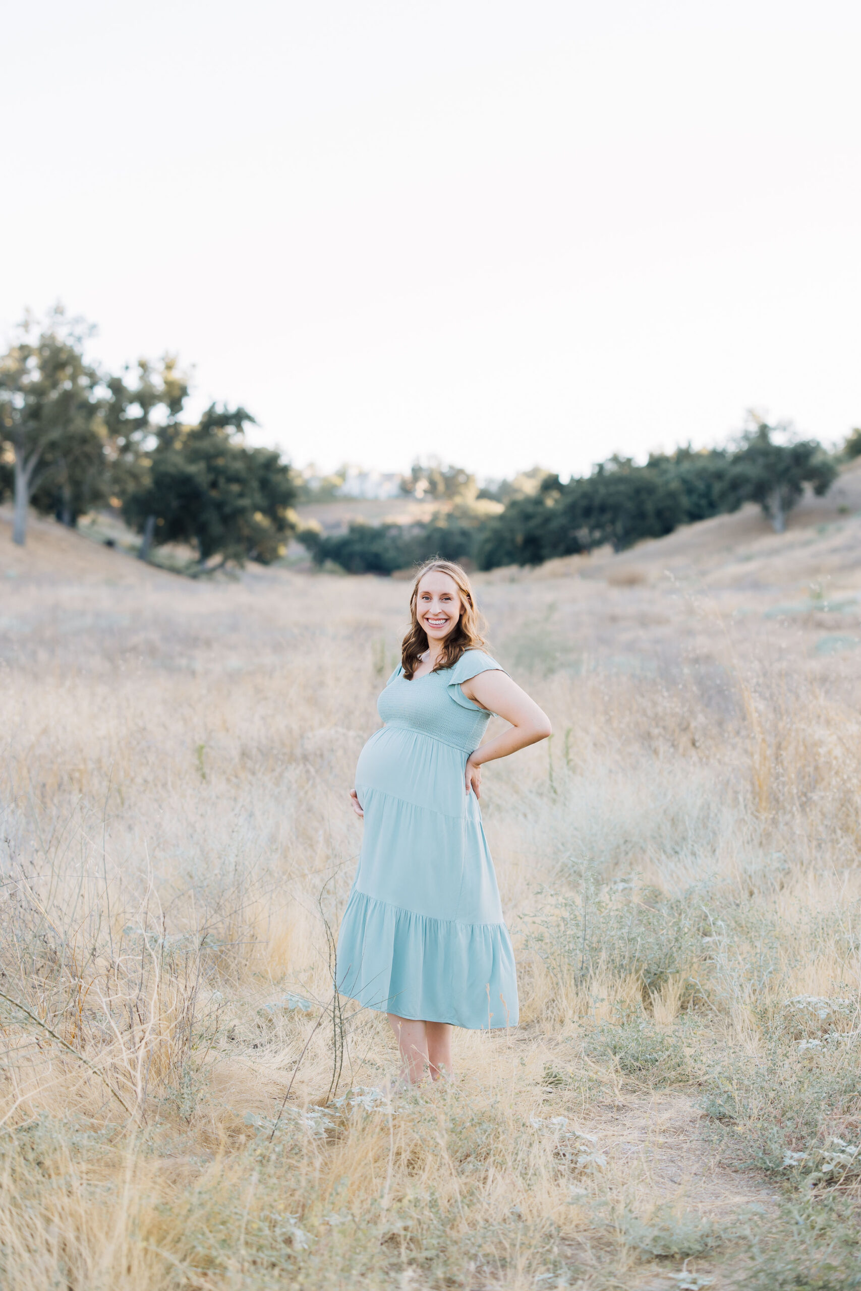 A proud mom to be stands in a field of tall golden grass in a blue maternity dress holding her bump before meeting a lactation consultant santa clarita