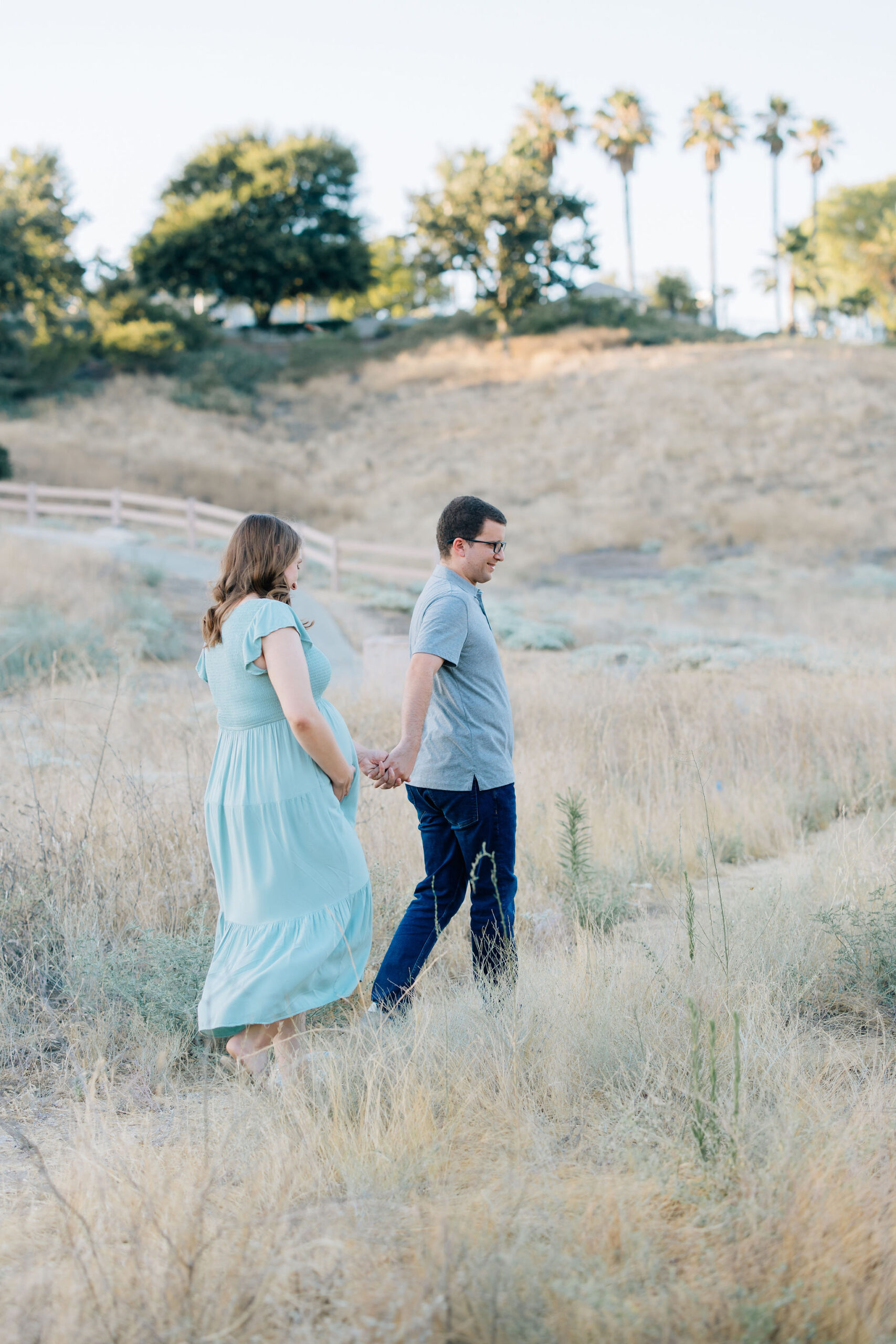 Happy expecting parents walk hand in hand through a park trail at sunset before meeting a lactation consultant santa clarita