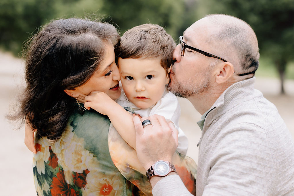 Happy parents kiss the head of their toddler son in a park