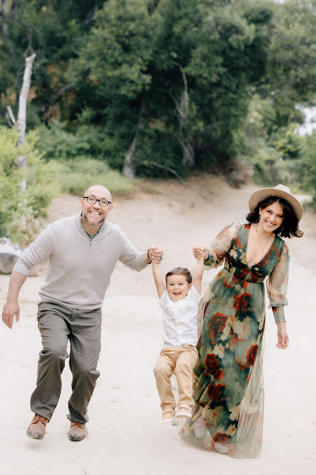 A happy toddler boy swings in mom and dad's hands in a park path as they all laugh before visiting pediatrician santa clarita