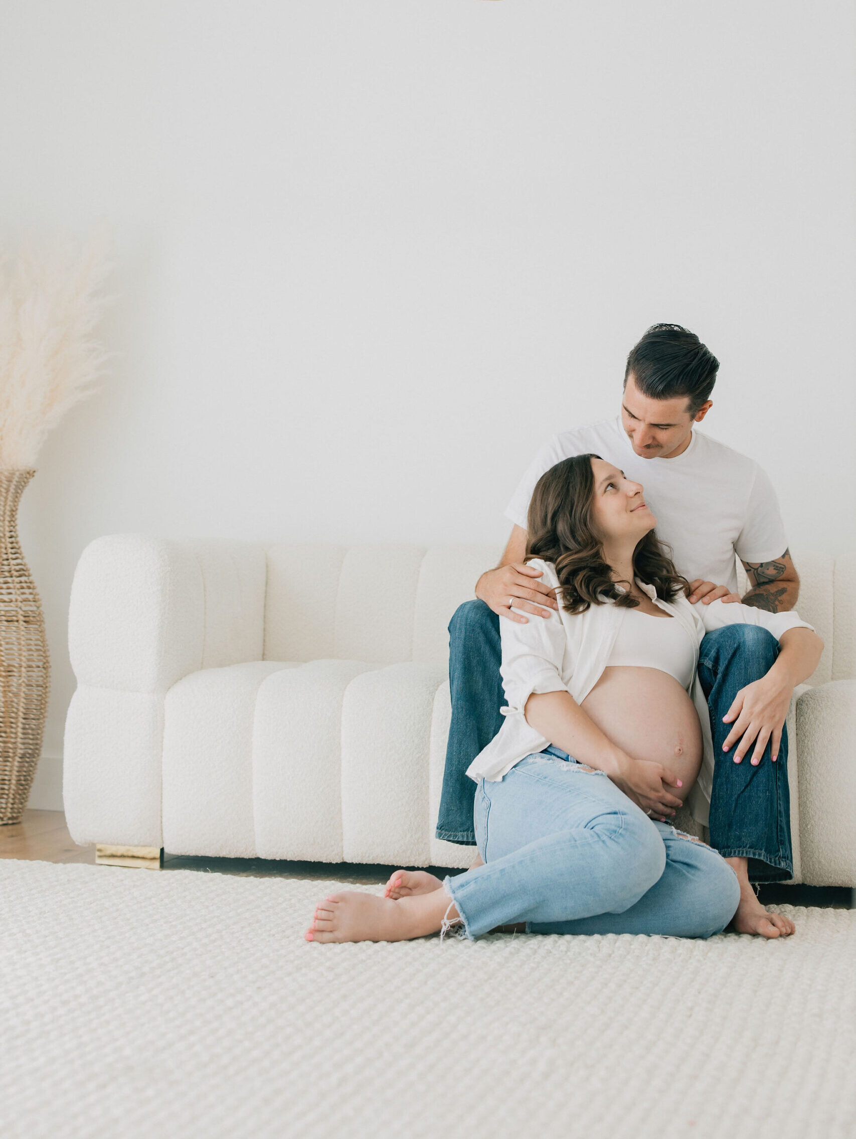 A happy expecting couple snuggles against a couch in a studio in jeans and white shirts while holding the bump