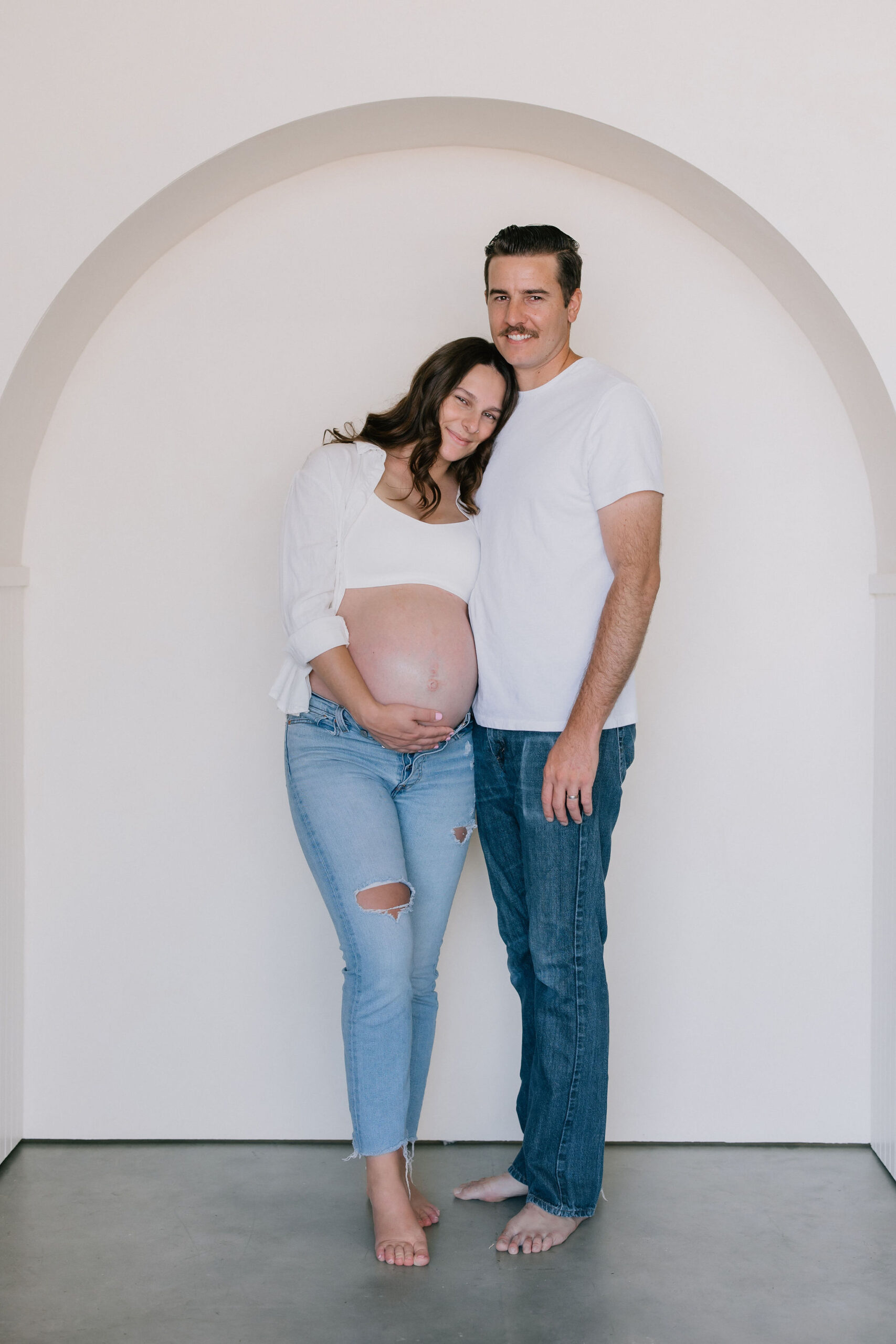 A pregnant woman in jeans and open white shirt leans and snuggles onto her husband as they stand in a white arch after visiting baby shower venues in santa clarita