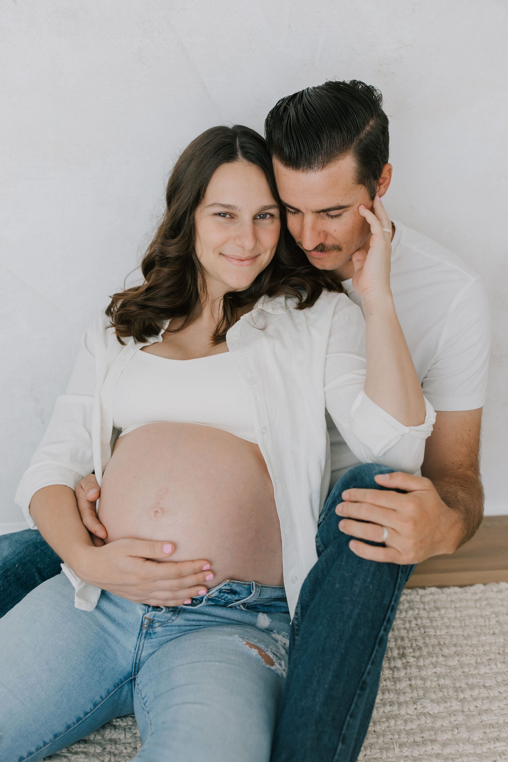 Happy expecting parents snuggle against a wall with the bump exposed in jeans and white shirts after visiting baby shower venues in santa clarita