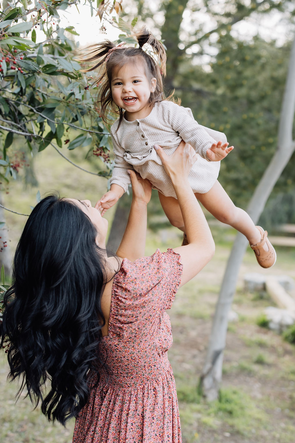 A giggling toddler girl in a white dress is lifted and tossed in the air by mom in a red dress