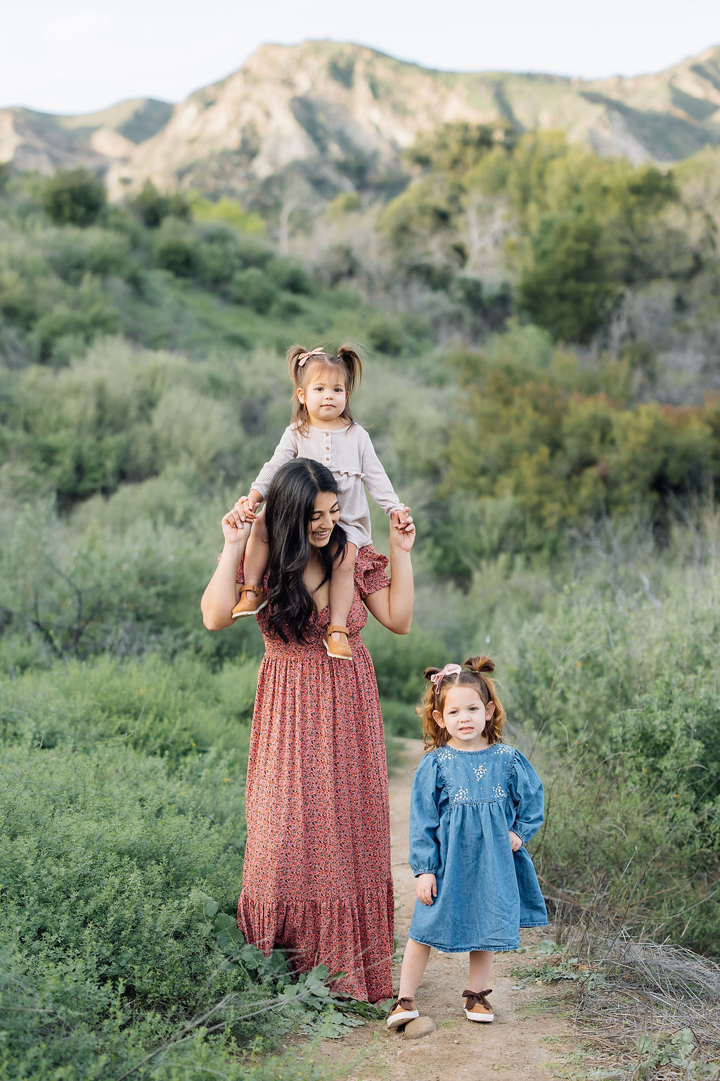 A toddler girl sits on mom's shoulders while older sister walks beside her in a mountain trail at sunset before finding christmas trees in santa clarita