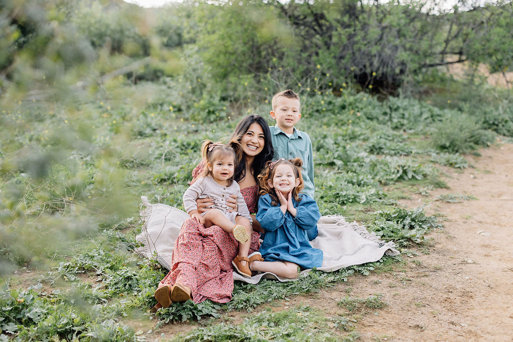 A happy mom sits on a picnic blanket in a park trail with her 3 happy toddlers around her before finding christmas trees in santa clarita