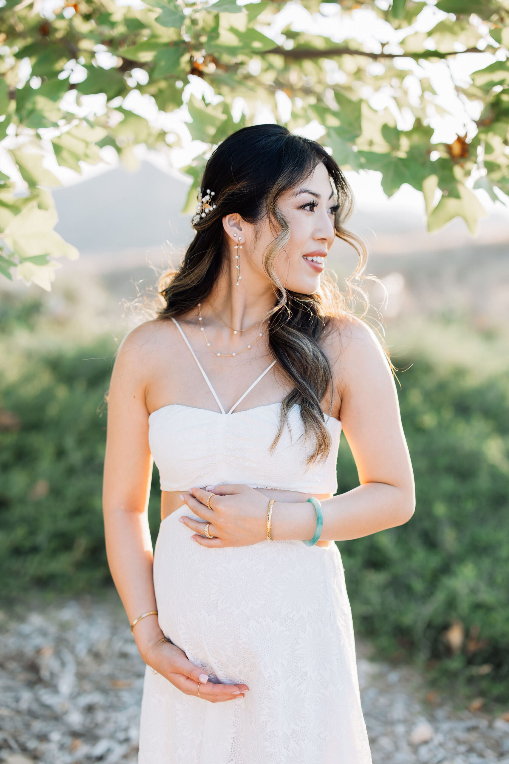 A mom to be in a white maternity gown smiles over her shoulder while standing under a tree at sunset after visiting an obgyn santa clarita