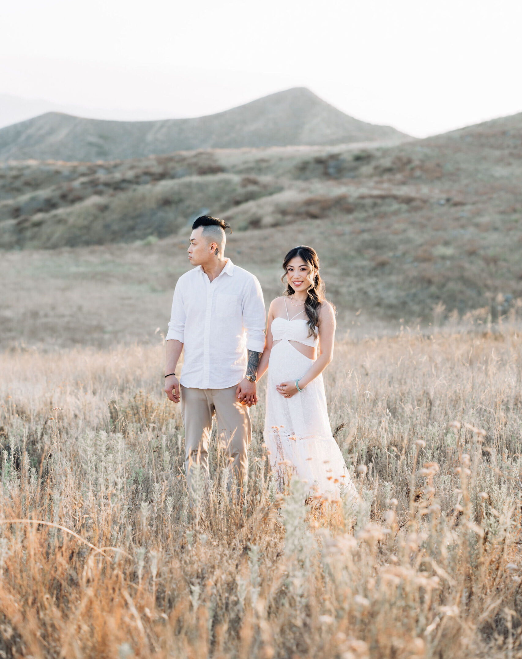 An expecting couple holds hands while exploring a field of tall golden grass at sunset in white clothes after finding an obgyn santa clarita