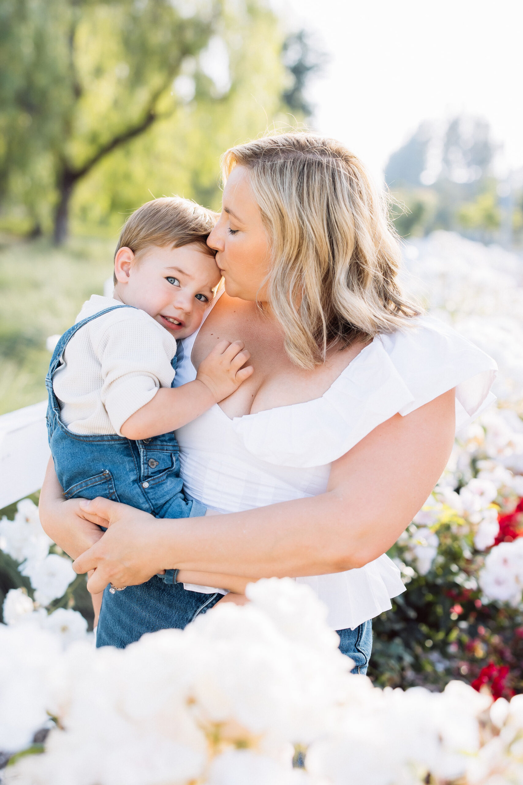 A toddler boy smiles while sitting on mom's hip and being kissed in a flower garden after visiting a pediatric dentist santa clarita