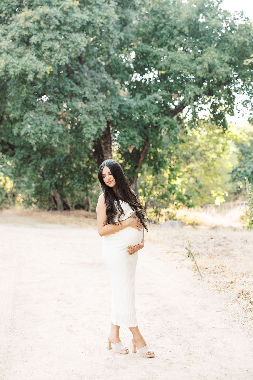 A smiling mother to be looks over her shoulder in a white maternity gown in a park path after visiting a prenatal chiropractor in santa clarita