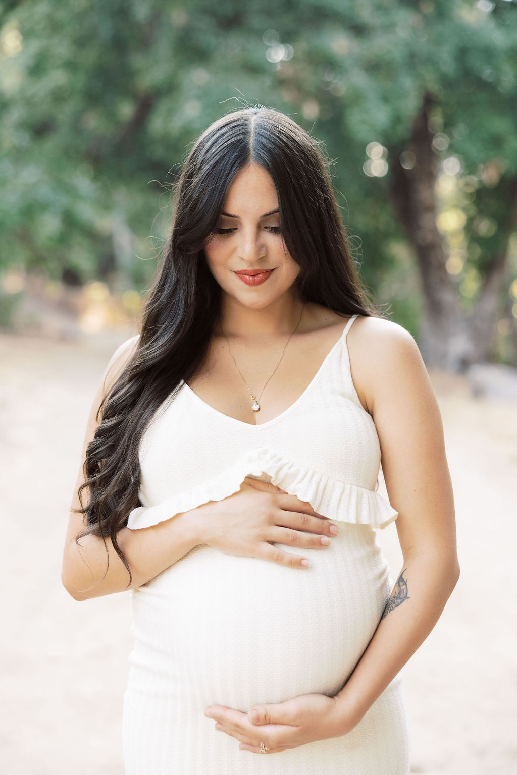 A mom to be in a white maternity dress stands in a park at sunset smiling down to her bump with her hands on it after visiting a prenatal chiropractor in santa clarita