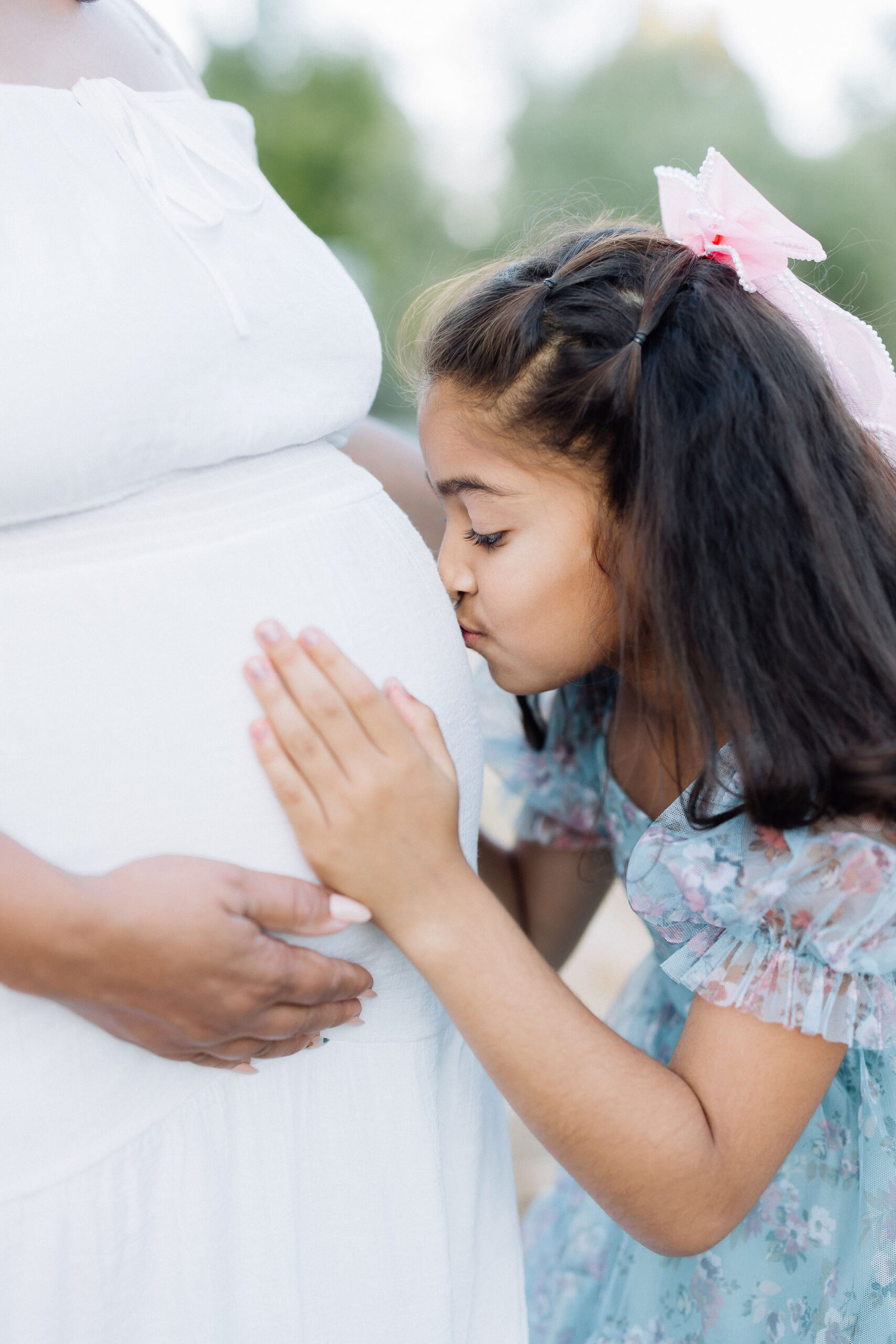 A young girl in a blue dress kisses mom's pregnant bump in a park