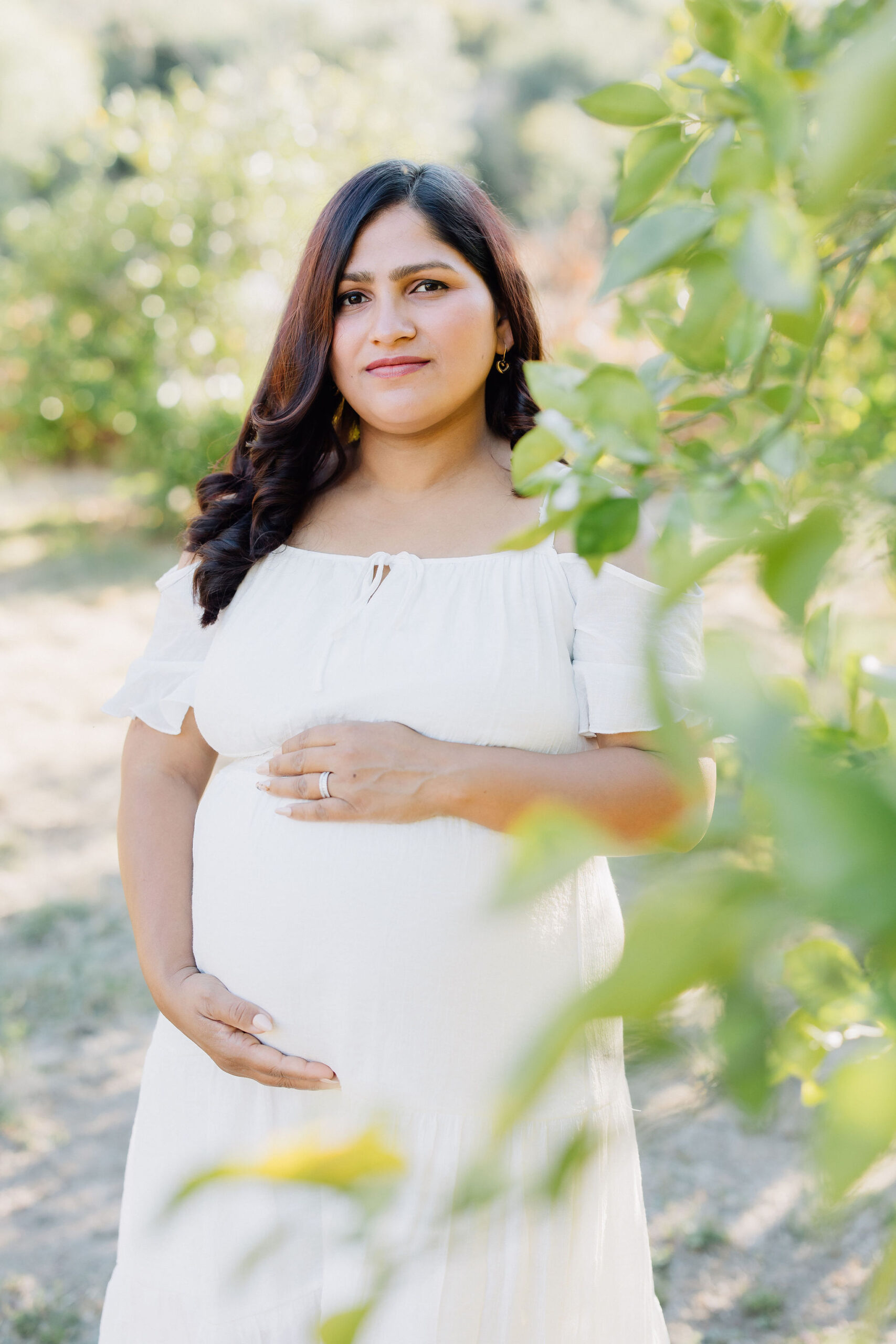 A happy mom to be stands in an orange grove in a white dress holding her bump after a prenatal massage in santa clarita