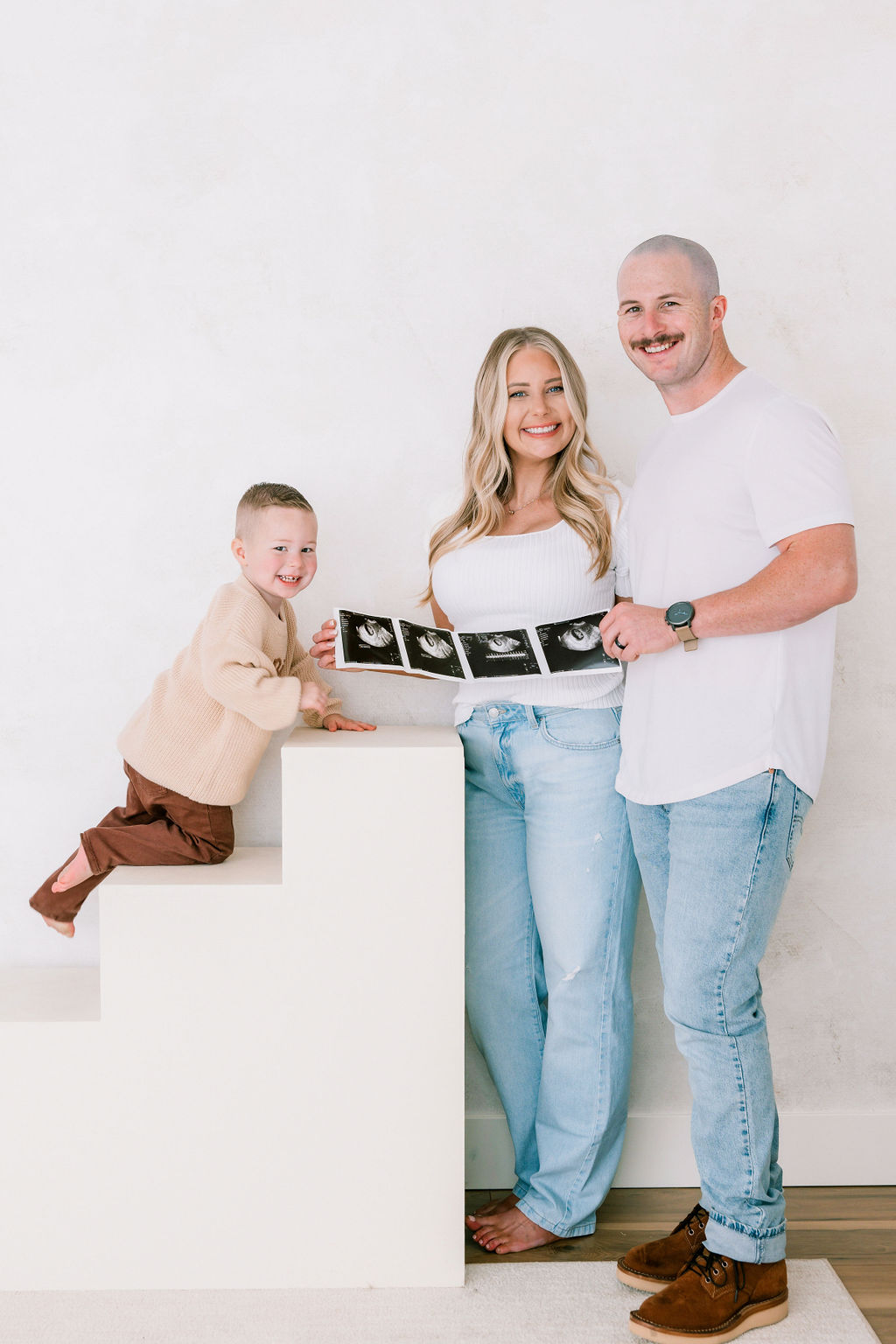 Happy expecting parents hold their sonogram as their toddler son climbs white stairs in a studio