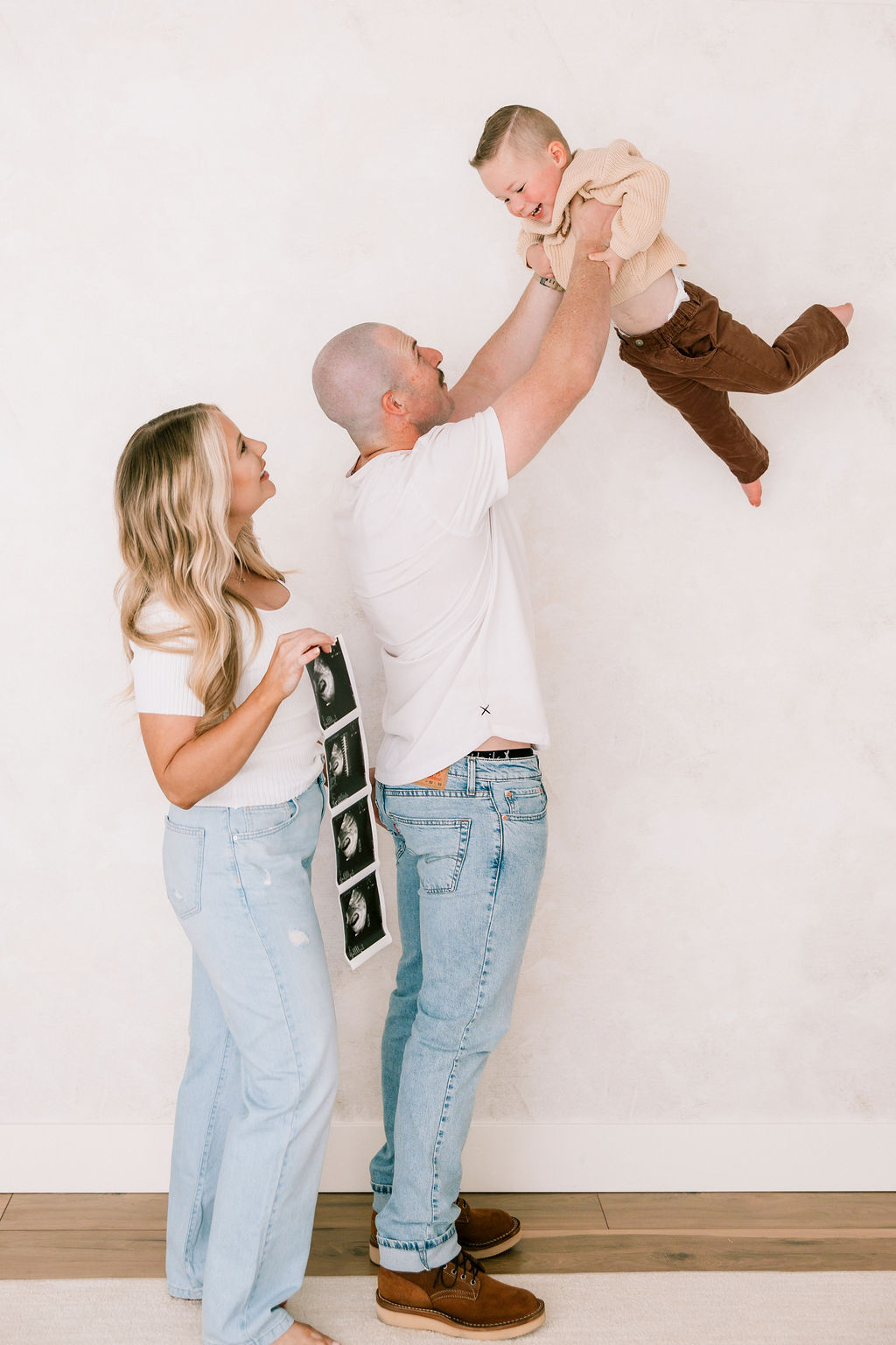 A happy toddler is lifted by dad in the air while standing in a studio with mom holding ultrasound scans before some prenatal yoga in santa clarita