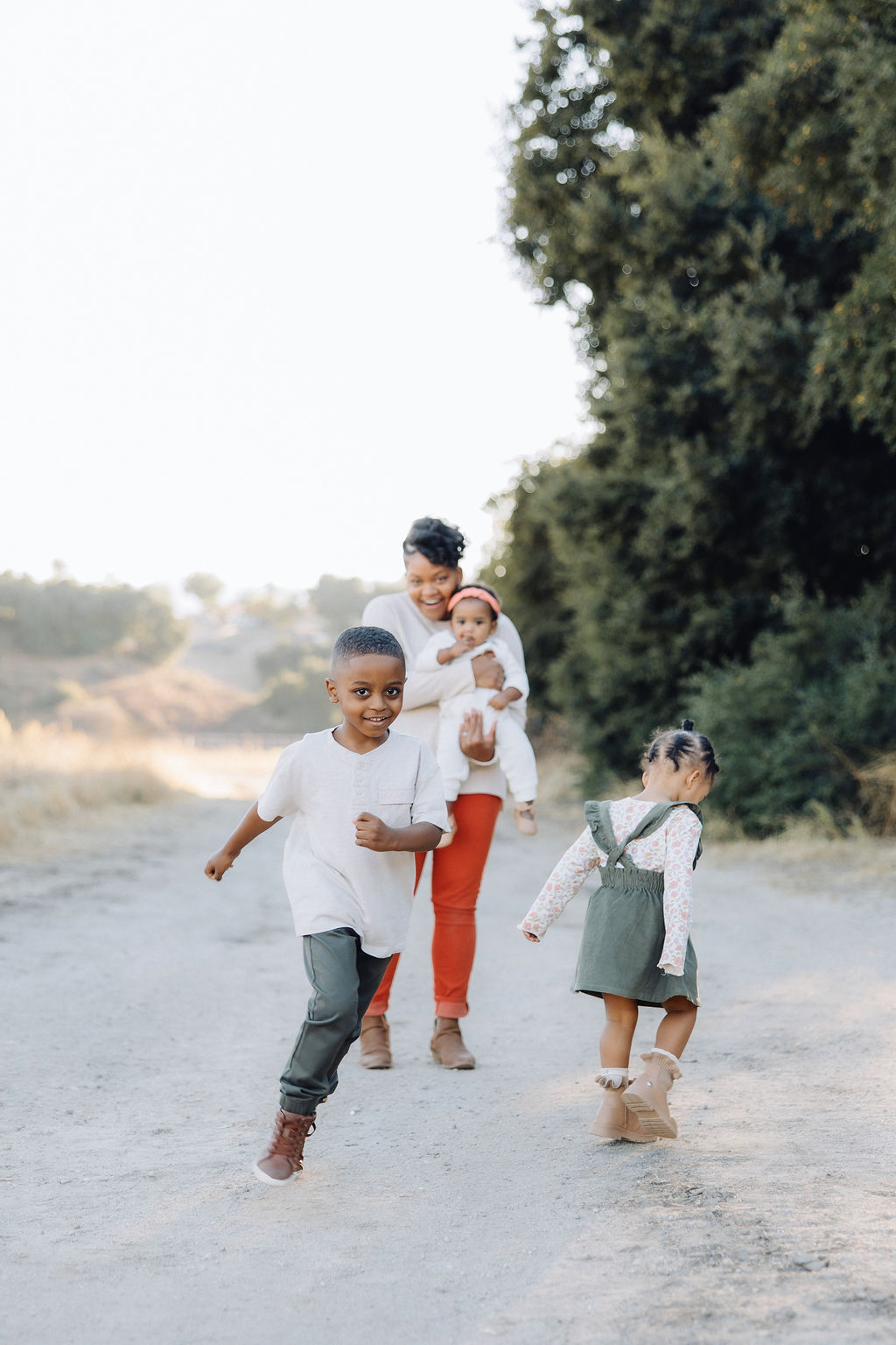A young boy and girl play and run in a park trail at sunset while mom watches with a big smile