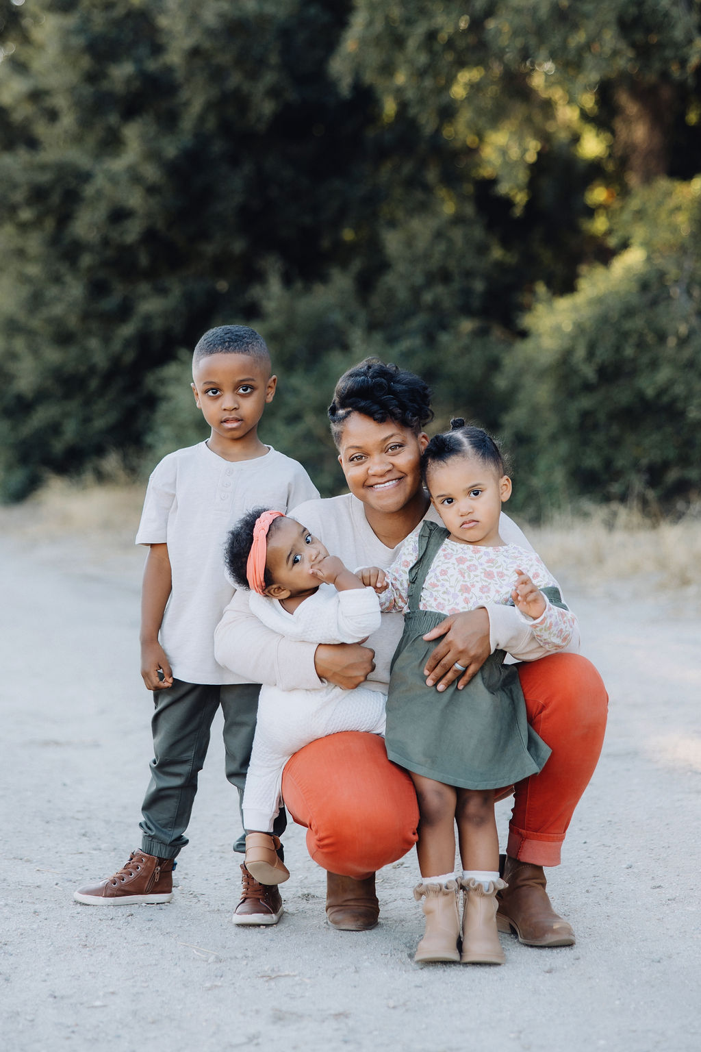 A happy mother squats in a park trail with her three toddlers in her arms in red pants after some parenting classes in Santa Clarita