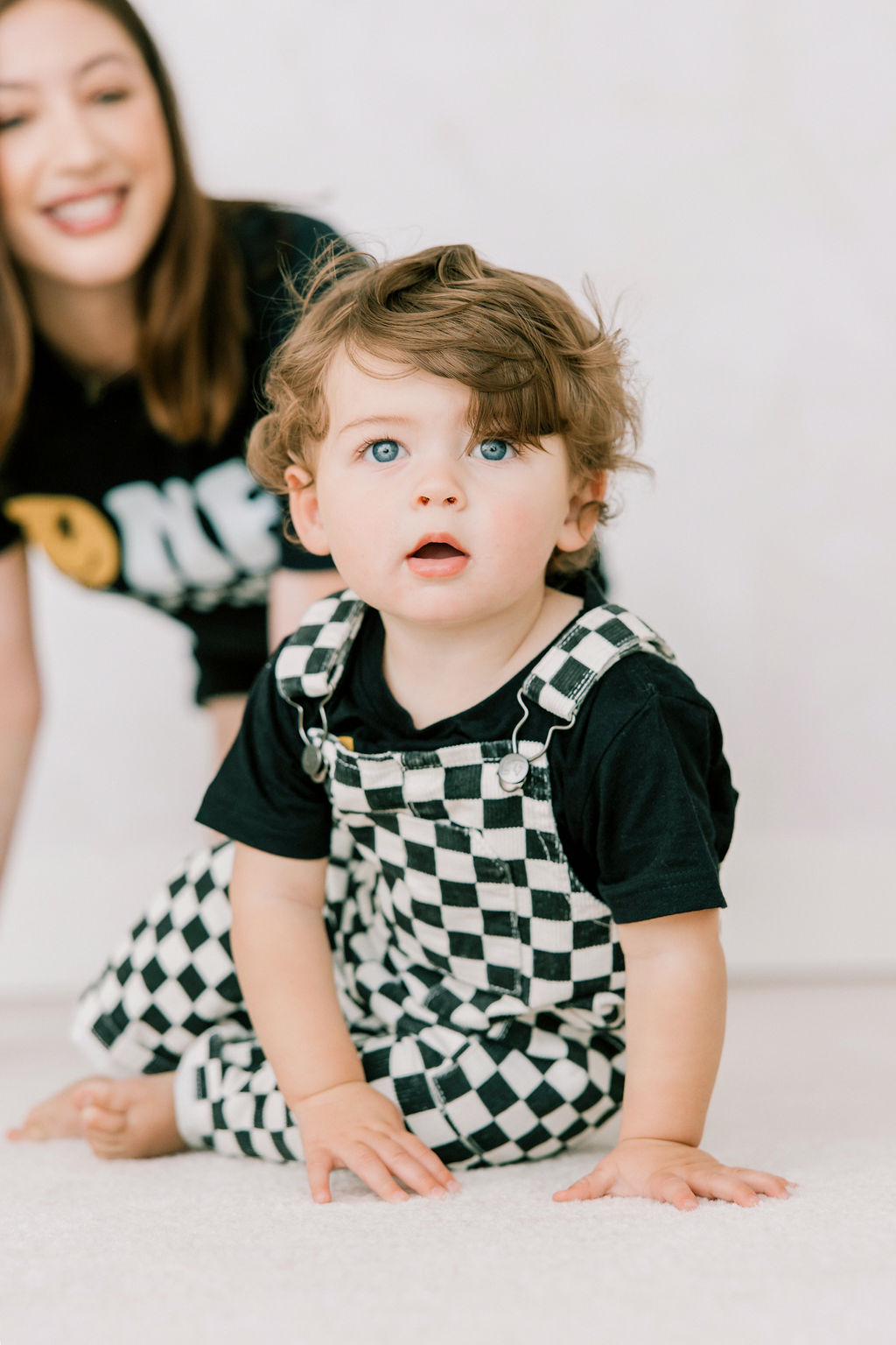 A happy mom watches as her one year old crawls on a carpet in a studio in checkerboard overalls