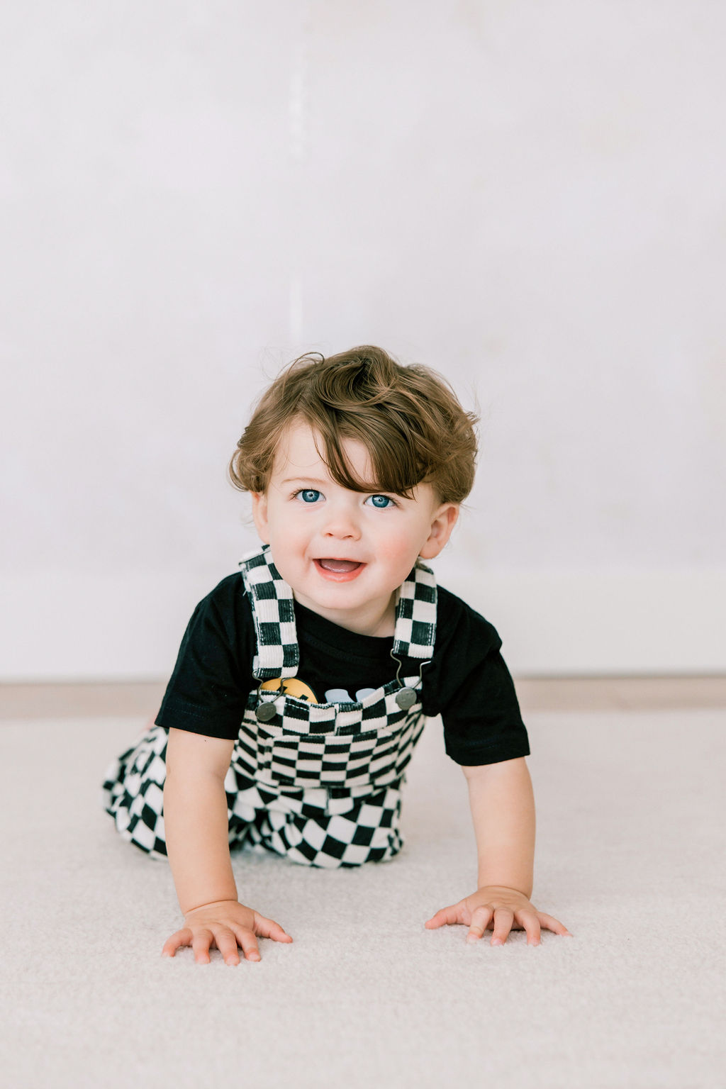 A baby in checker board overalls and black shirt crawls through a studio smiling before visiting preschools in Santa Clarita