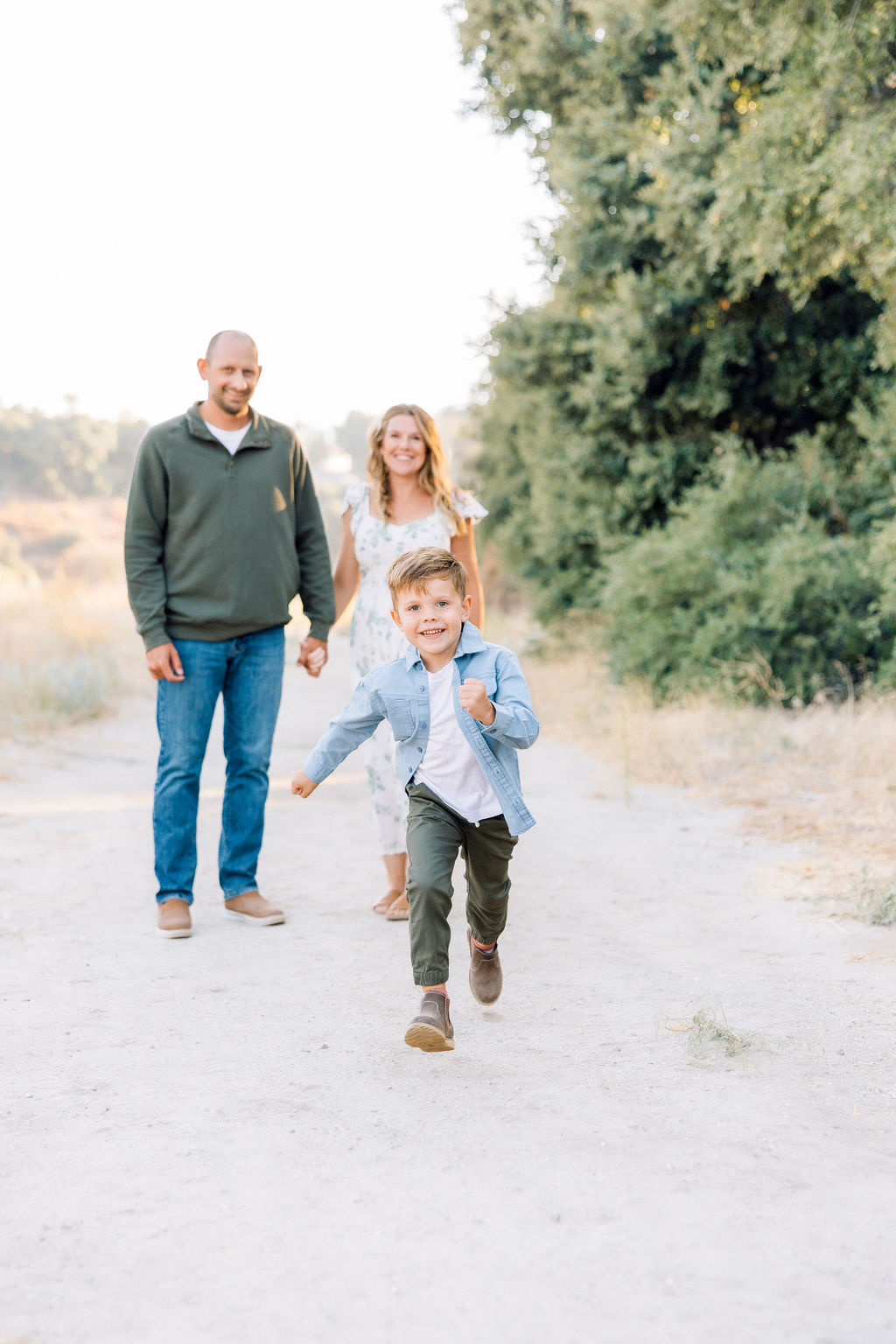 A young boy in a blue shirt and green pants runs in a park trail as mom and dad watch