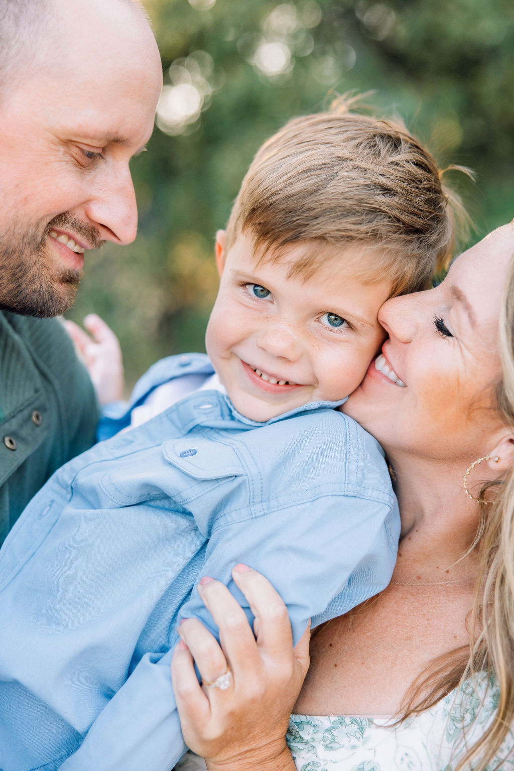 A mom and dad snuggle their toddler son in a blue shirt in a park as he smiles after some swim lessons in Santa Clarita