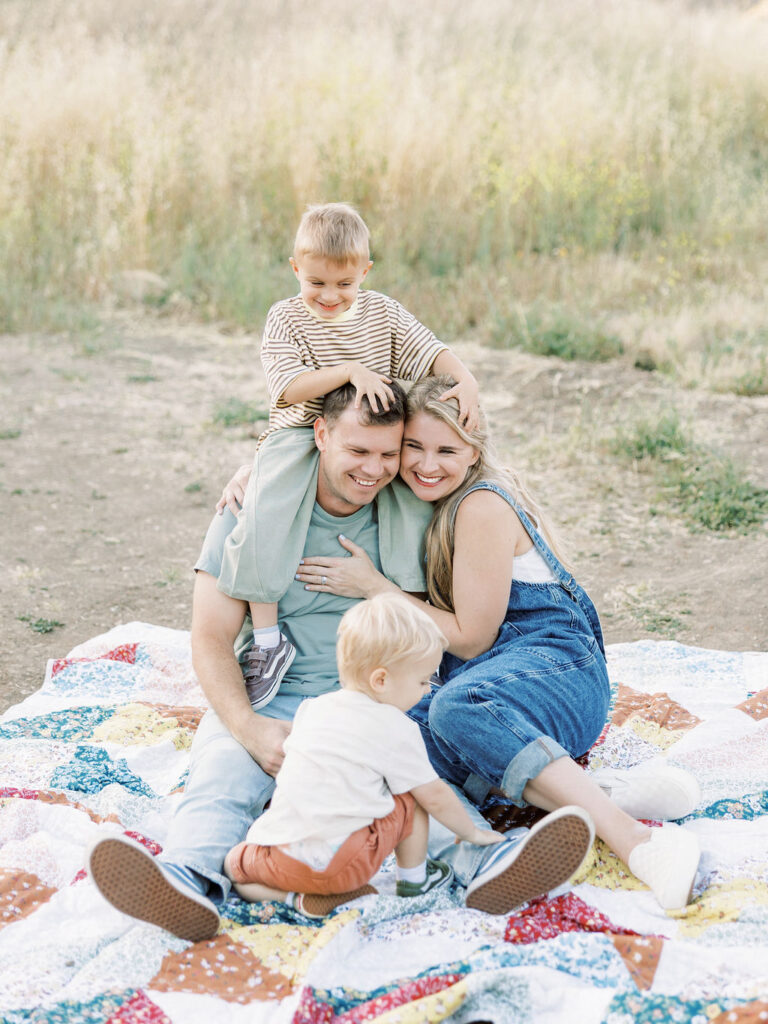 Family of four cuddling together on a quilt while one toddler sits on dad’s shoulders during playful family photos with toddlers in Santa Clarita.