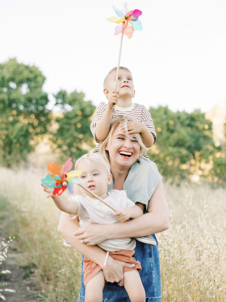 Mom laughing while holding one toddler and another sits on her shoulders with colorful pinwheels during playful family photos with toddlers in Santa Clarita.
