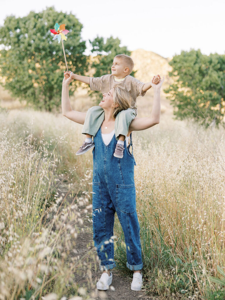 Mom carrying her toddler on her shoulders while he holds a colorful pinwheel during playful family photos with toddlers.