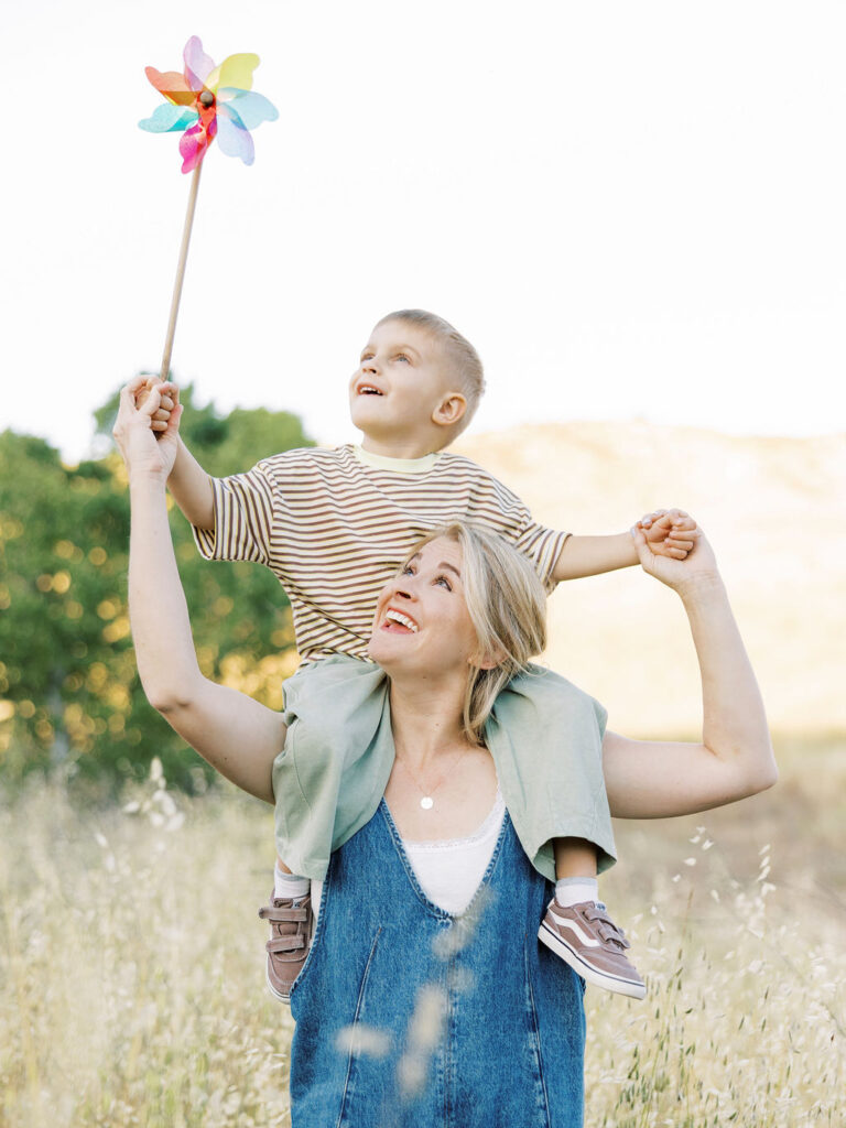 Mom carrying her toddler on her shoulders while he holds a colorful pinwheel during a playful Santa Clarita family photography session.