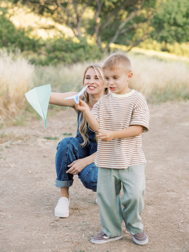Mom crouching behind her toddler as he plays with a paper airplane during a candid Santa Clarita family photography session.