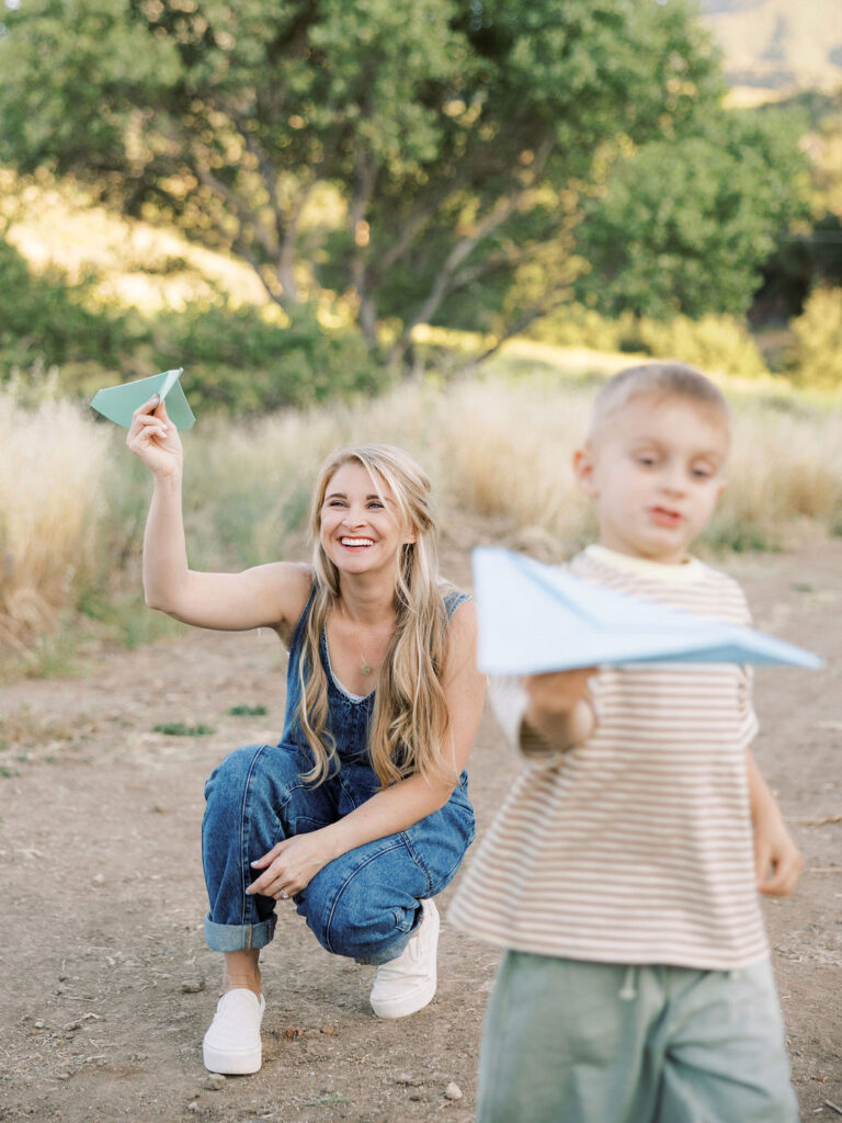 Mom smiling behind her toddler as he plays with a paper airplane during a candid Santa Clarita family photo session.
