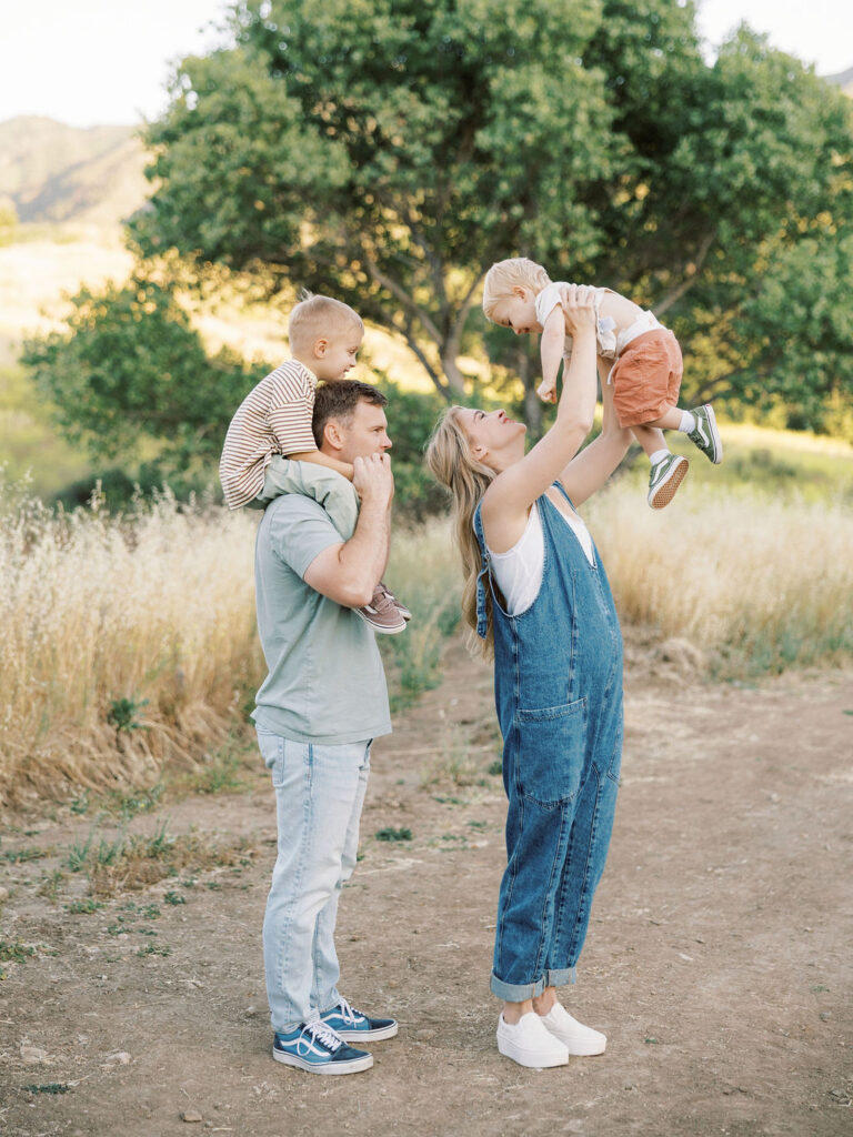 Dad carrying one toddler on his shoulders while mom lifts their younger child into the air during playful family photos with toddlers in Santa Clarita.