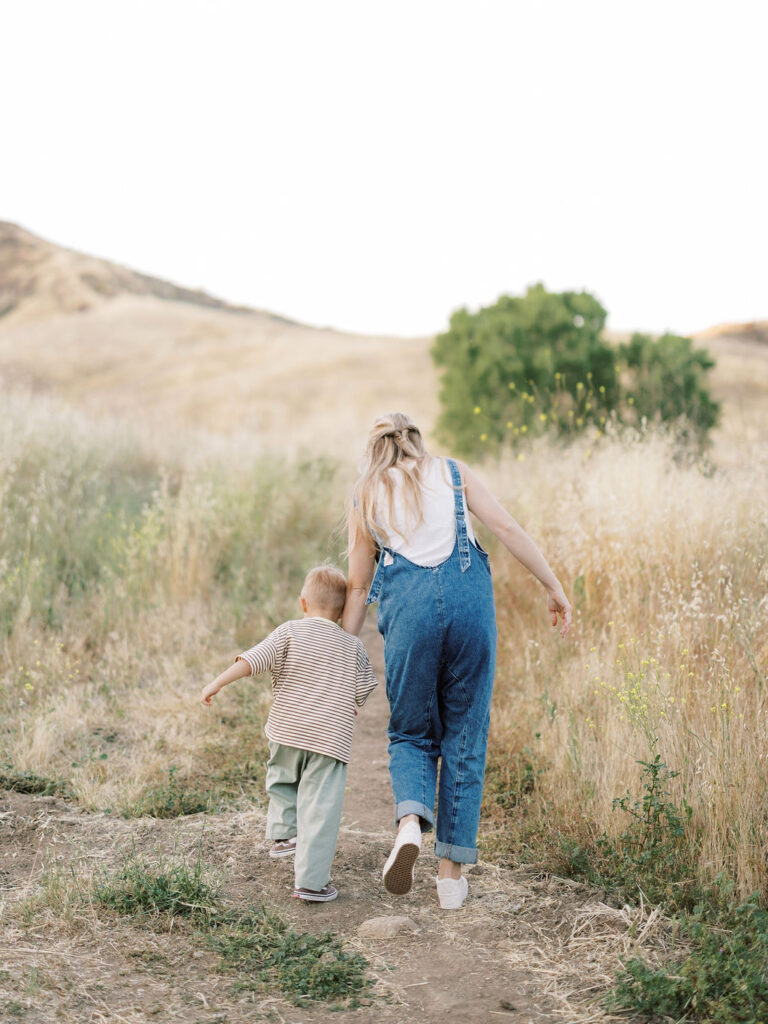 Mom and toddler boy walking through tall grass during natural family photos in Santa Clarita