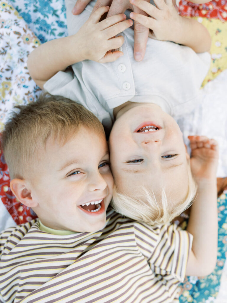 Two young siblings lying together on a colorful quilt, laughing and cuddling during playful family photos with toddlers in Santa Clarita.